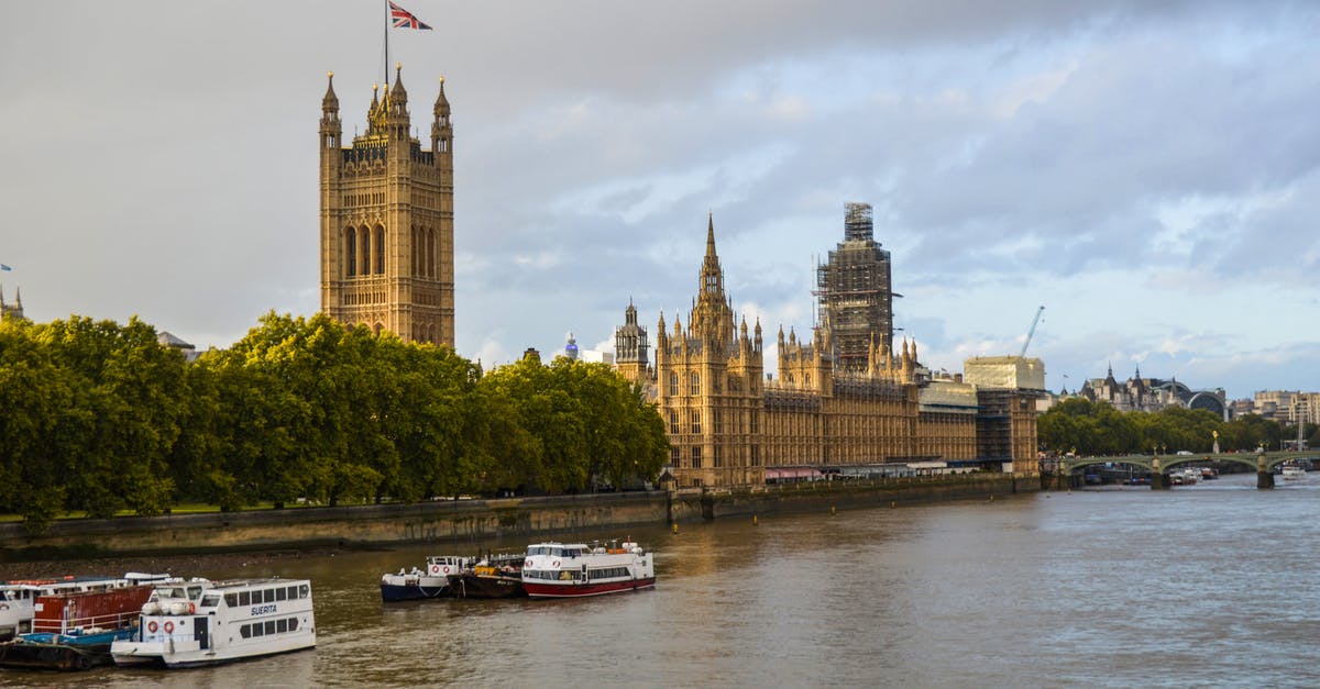 River Thames Source and Destination Points - Exterior of aged building of the Houses of Parliament on bushy coast near river with vessels under cloudy sky of London