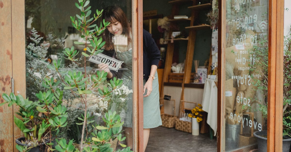 Schengen Single entry Business Visa to Multi entry - Positive young Asian female florist in apron standing near entrance and turning signboard on glass wall while working in modern floristry shop