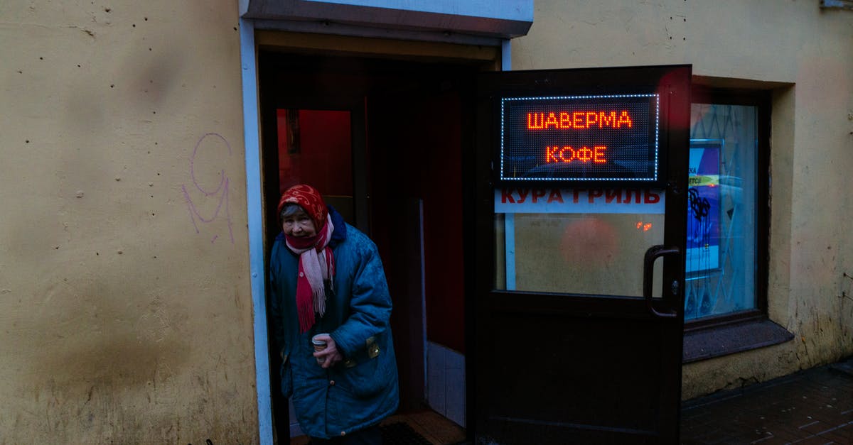 Shop opening hours in Russia - Senior gloomy female in shawl and warm jacket with cup of coffee leaning on entrance of bistro and looking at camera