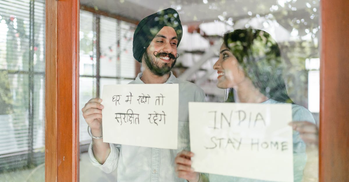 Short Stay Schengen Visa for non Eu spouse - Through glass of young Indian couple standing against window and looking at each other while demonstrating sheets of paper with India stay home inscription in English and Hindi