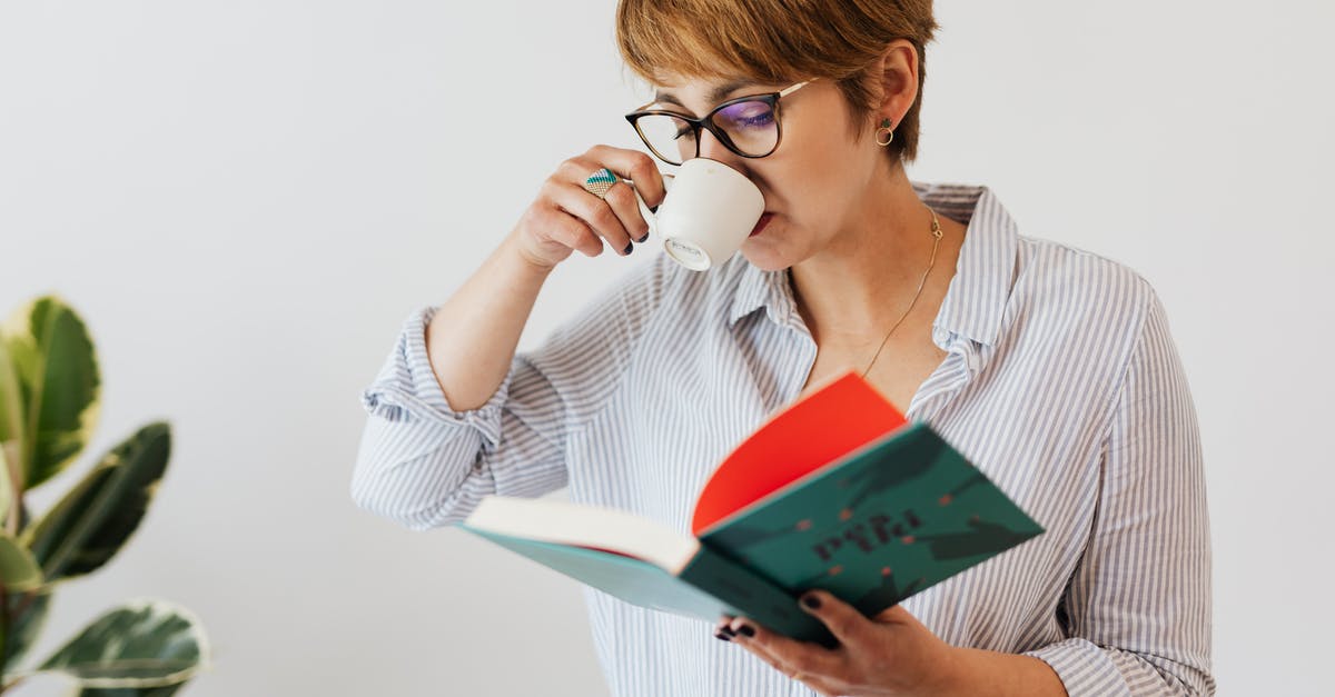 Short-term study visa UK - Crop pensive female in eyeglasses and casual wear reading interesting book in hardcover and drinking hit aromatic coffee from white ceramic cup against white wall and green plant