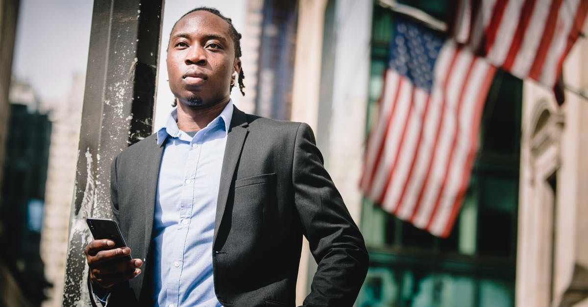 Should I always use the same nationality to enter the USA? - Low angle of serious well dressed ethnic male manager leaning on metal pillar on street and using cellphone against government building with hanging American flags