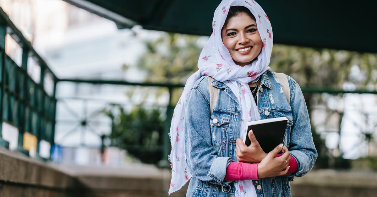 Should I cover up my tattoo when visiting the Middle East? - Content ethnic female in headscarf and denim coat with notebook looking at camera with toothy smile