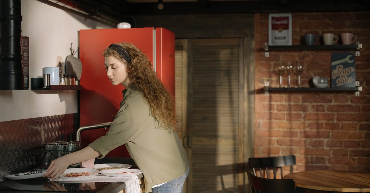 Should I wash dishes before leaving a serviced apartment? - Woman in Gray Long Sleeve Shirt and Blue Denim Jeans Standing Beside Kitchen Counter