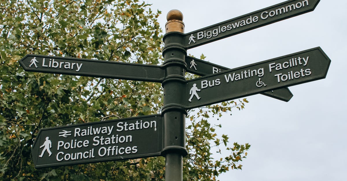 Show the time at milestones for a car travel route - From below poled signpost placed near lush green tree and showing direction to city facilities in daylight