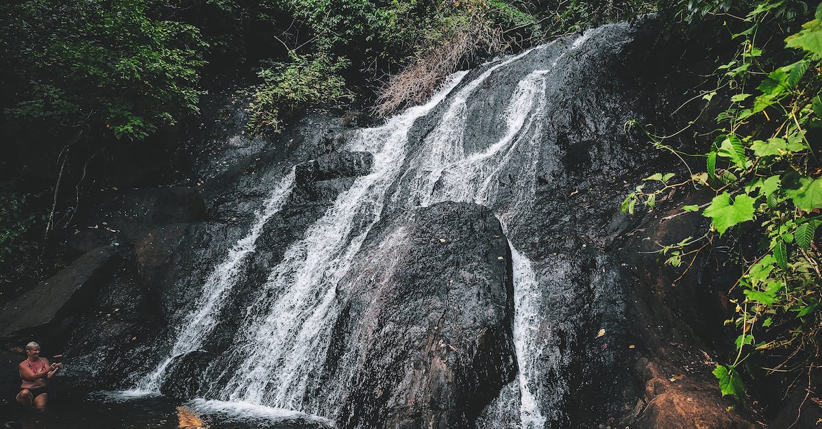 Shower for a traveler passing through Delhi - Unrecognizable travelers chilling in warm pond formed by waterfall flowing through rough stony cliff in lush forest