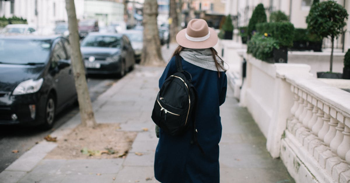 Sightseeing in the Dakotas without a car - Back view of unrecognizable stylish female tourist with backpack in trendy coat and hat walking along pavement near parked cars on street and modern buildings during daytime