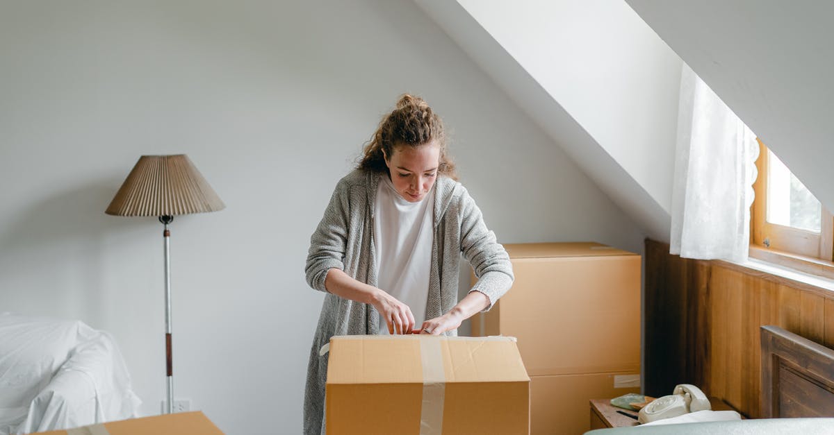 Stapled old and new passports - Slim female in cardigan unpacking cardboard box put on bed close to suitcase while standing near window in attic style room in daylight