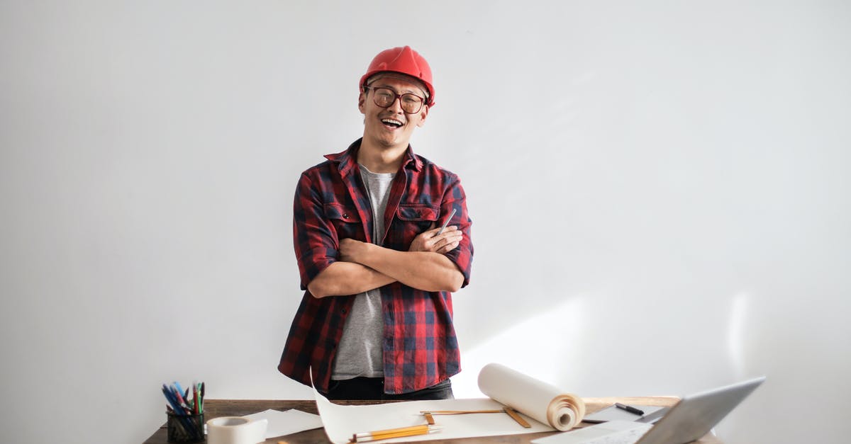 Strategy to improve seats on a flight? - Smiling casual man in hardhat and glasses holding arms crossed looking at camera while standing at desk with paper draft and stationery