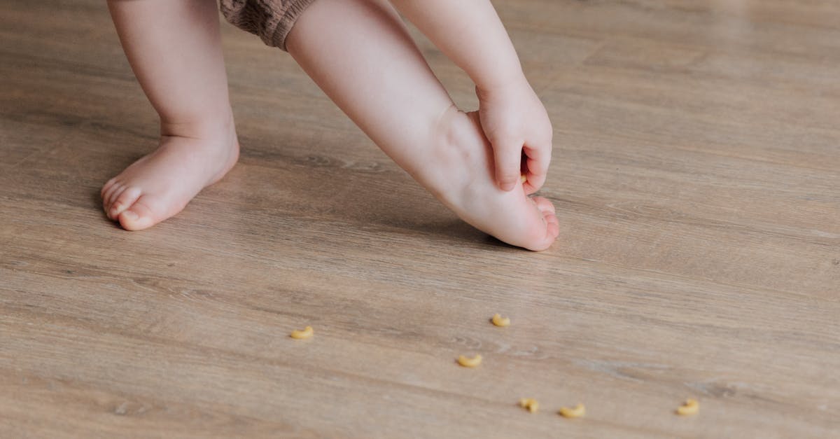 Stuck in another country? - Crop faceless toddler standing barefoot on floor and trying to remove stuck pasta from foot while playing and developing fine motor skills at home