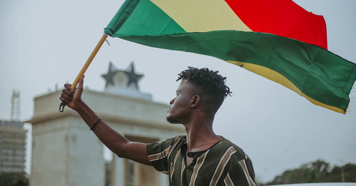 Stuck in Ghana with $3000 [duplicate] - African male with dreadlocks raising flag of Ghana country with colorful stripes while looking away in town