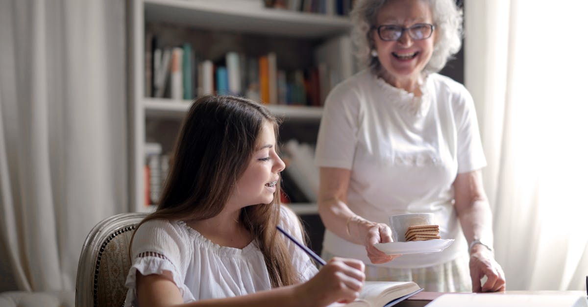 Suggestions for 15 year old ( child / teen ) travelling alone - Cheerful grandmother in glasses and casual clothes smiling at camera while giving pastry on plate and mug of tasty beverage to joyful teenager sitting at table with books and exercise book and study in light cozy living room