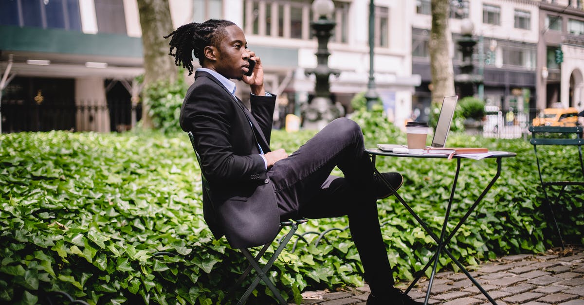 Surfing in Sri Lanka in August - Side view serious African American businessman in black formal wear sitting at table in city park with legs crossed and having conversation via modern smartphone