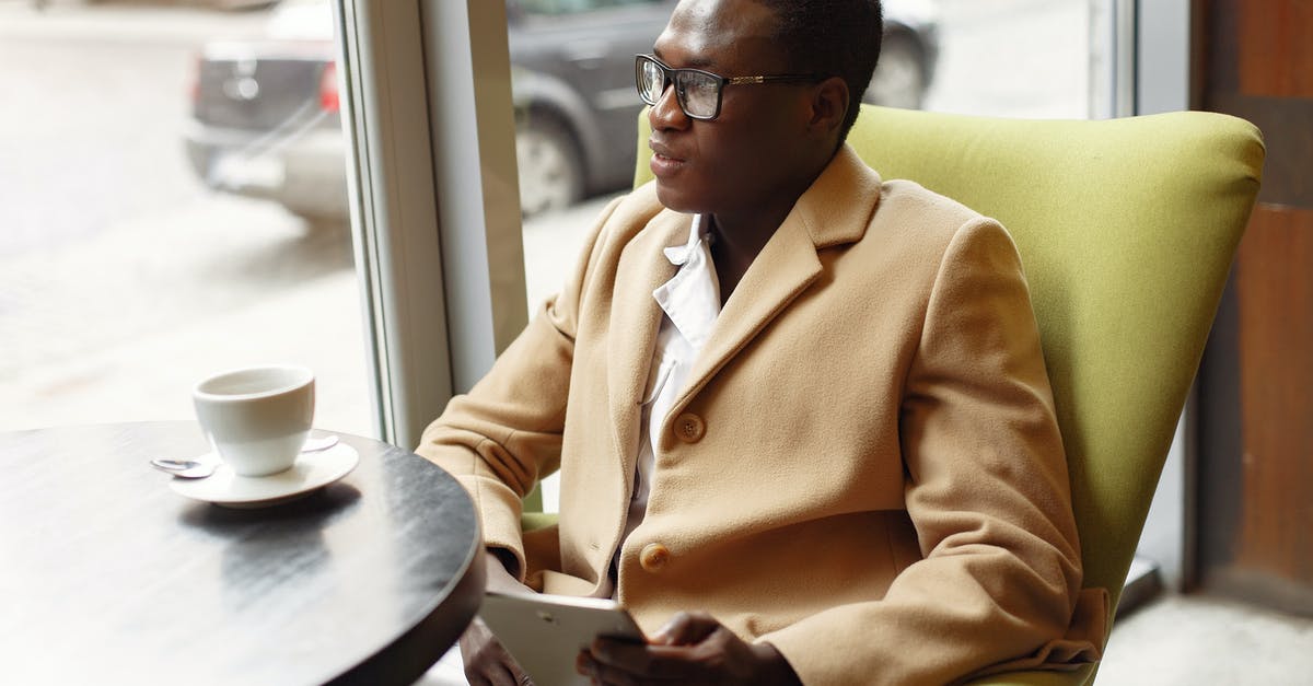 Surfing in Sri Lanka in August - Serious African American male in trendy formal suit and eyeglasses sitting on cozy chair in cafe with cup of coffee and browsing tablet