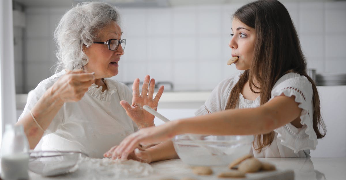 Teen child traveling with family member within US - Calm senior woman and teenage girl in casual clothes looking at each other and talking while eating cookies and cooking pastry in contemporary kitchen at home