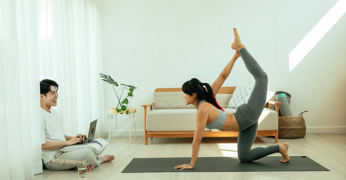 Ticket for Universal Studios LA - Online or Physical - Side view of young barefoot female in sportswear performing Tiger pose on mat during yoga session while Asian smiling male sitting crossed legged on floor and using netbook