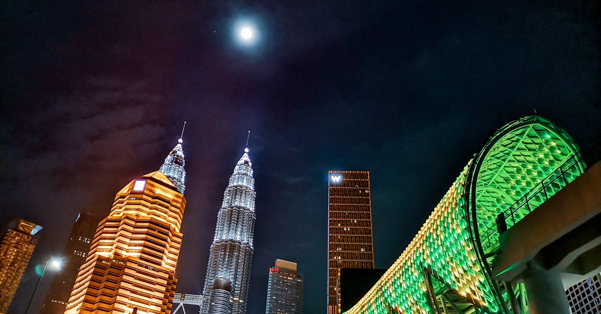Time to get to KUL from center of Kuala Lumpur - From below exterior of contemporary Petronas Twin Towers located amidst urban city towers and pedestrian overpass illuminated by electrical lights at night