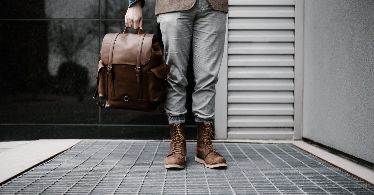 Toiletries in hand bag while travelling - Photo of Man in Brown Blazer,Gray Pants, and Brown Boots Holding Brown Leather Bag Standing Outside Building