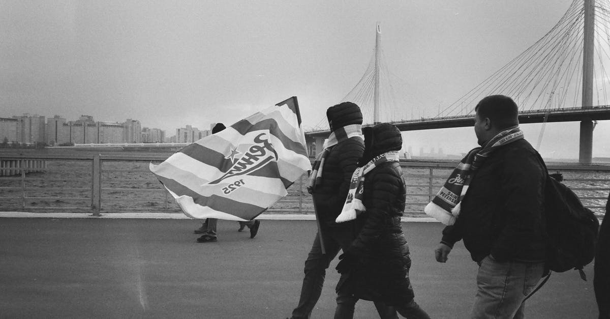 Tourist Schengen visa dates do not match itinerary - Black and white side view of people in warm clothes with flag walking along river embankment against modern bridge