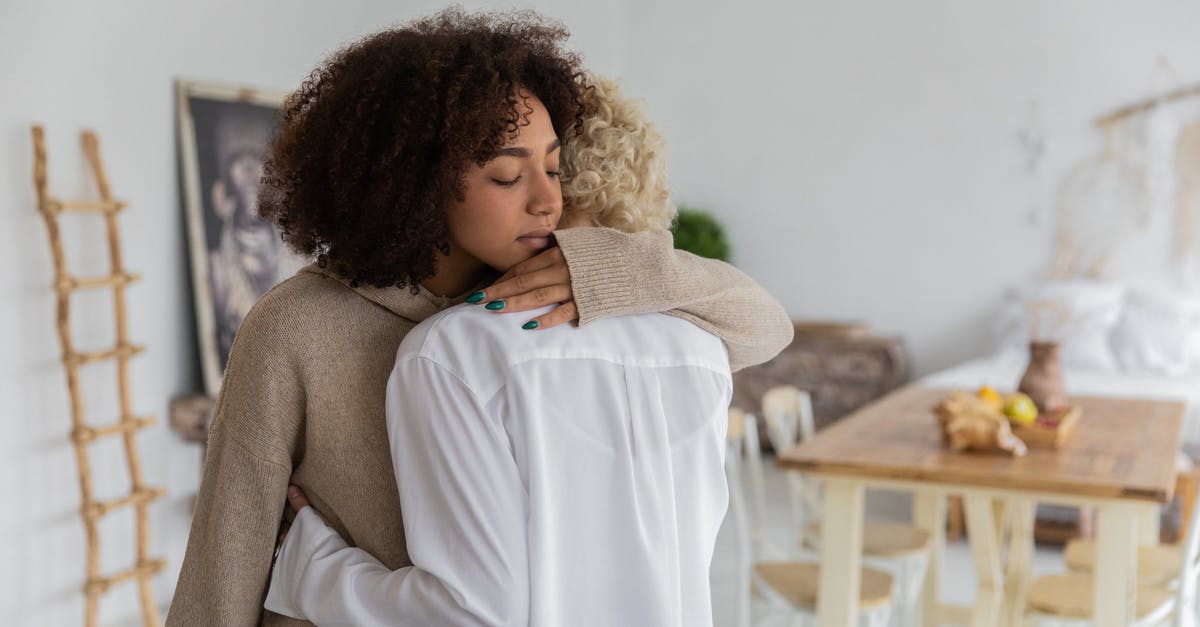 Transfer time at Newark domestic to domestic different carriers - African American lady hugging unrecognizable female blond friend during meeting in cozy light room with table and bed on blurred background