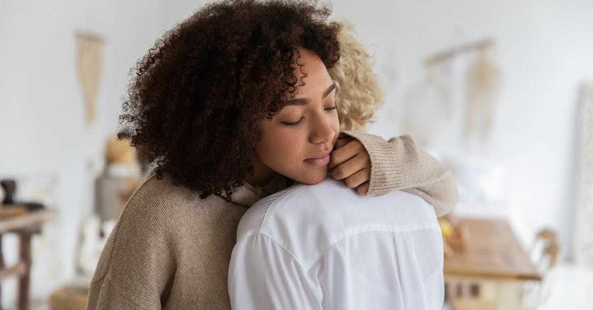 Transfer time at Newark domestic to domestic different carriers - African American woman embracing female anonymous friend while standing in cozy light room with wooden table and decorations on wall with blurred background