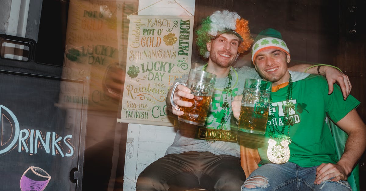 Transit through UK with Irish visa - Through glass wall view of cheerful best male friends with beer embracing while looking at camera during Feast of Saint Patrick in pub