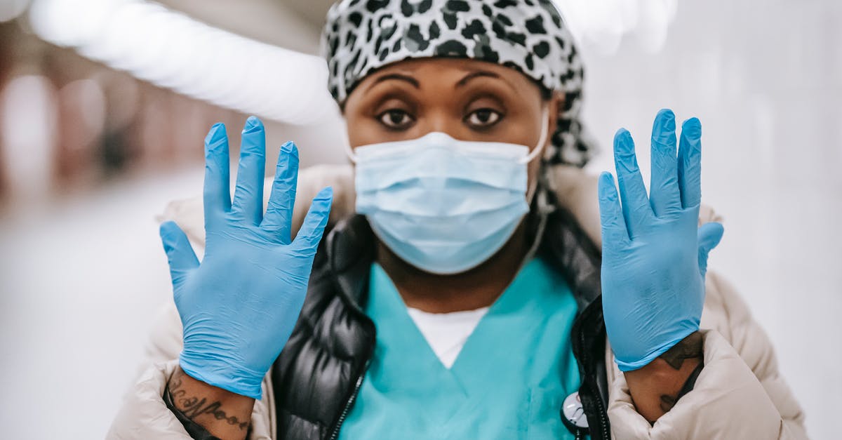 Travel advice back to UK from Jamaica after covid-19 outbreak - Pensive adult African American nurse wearing uniform warm clothes and face mask showing hands in blue latex gloves while standing in public hallway and looking at camera