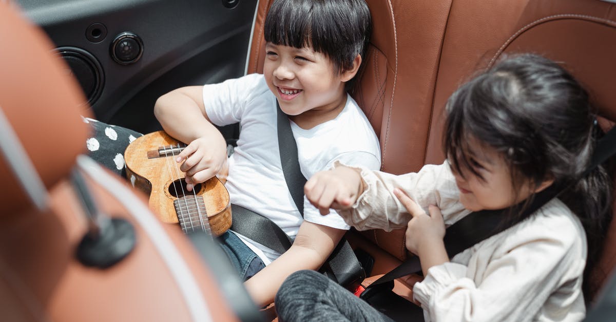 Travel from the UAE to Lithuania by car - From above smiling ethnic boy and girl in casual outfits sitting fastened in passenger seats with ukulele during road trip together