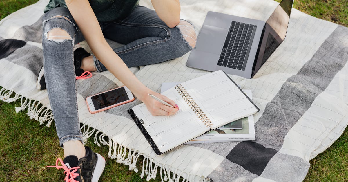 Travel planner app/website to plan by attractions/beaches/zoo/amusement park/pubs/Uber - From above crop anonymous female student in casual outfit writing in planner while sitting on warm blanket with smartphone and laptop on green park lawn