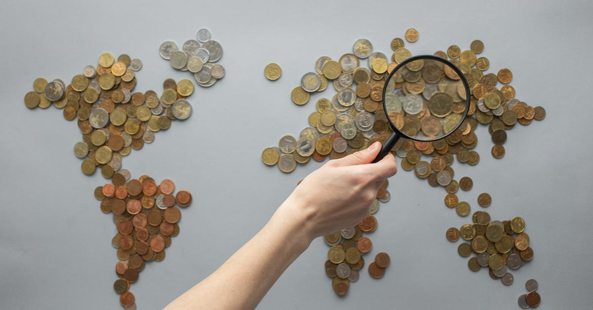 Travel to England from Argentina: how much money for a 17 day visit? - Top view of crop unrecognizable traveler with magnifying glass standing over world map made of various coins on gray background
