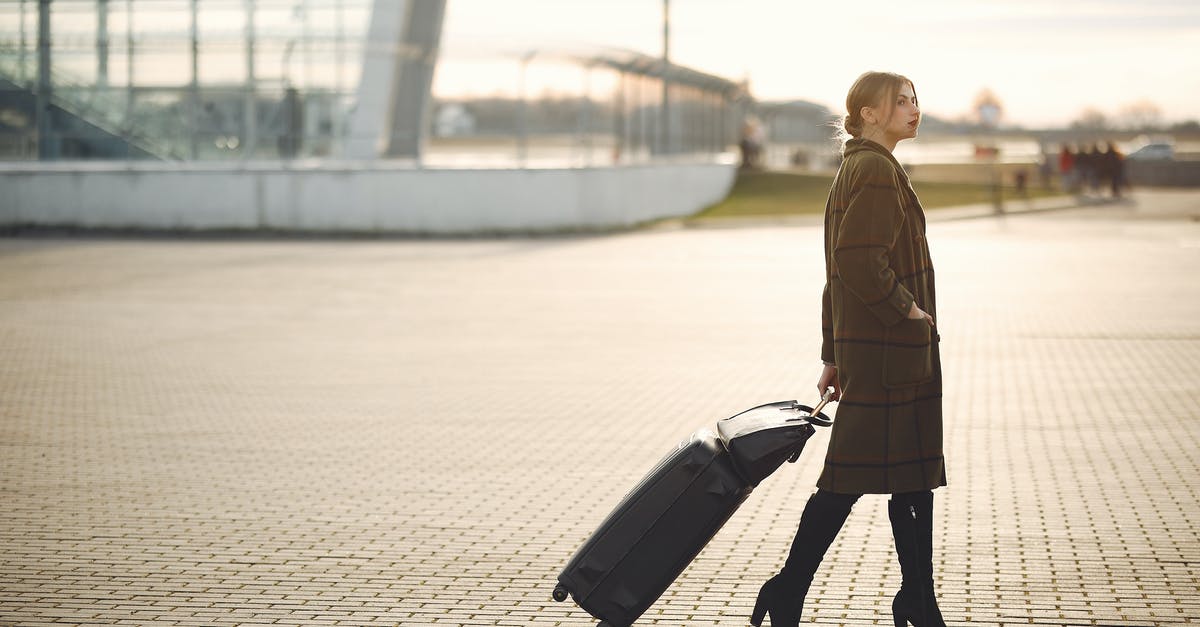 Traveling from Portoroz, Slovenia to Ljubljana airport on Sunday morning - Side view of full length young female traveler in trendy plaid brown coat and high boots with suitcase walking on pavement near modern building on city street in morning Traveling from Portoroz, Slovenia to Ljubljana airport on Sunday morning - Side view of full length young female traveler in trendy plaid brown coat and high boots with suitcase walking on pavement near modern building on city street in morning