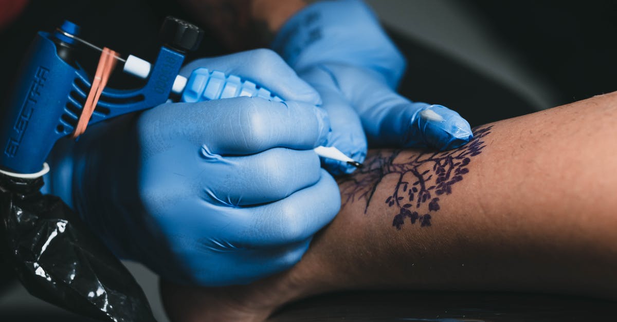 Traveling to Korea with special entry procedure because of COVID-19 - From above of faceless tattooist in blue gloves applying tree tattoo with ink and professional machine on leg of anonymous client in salon