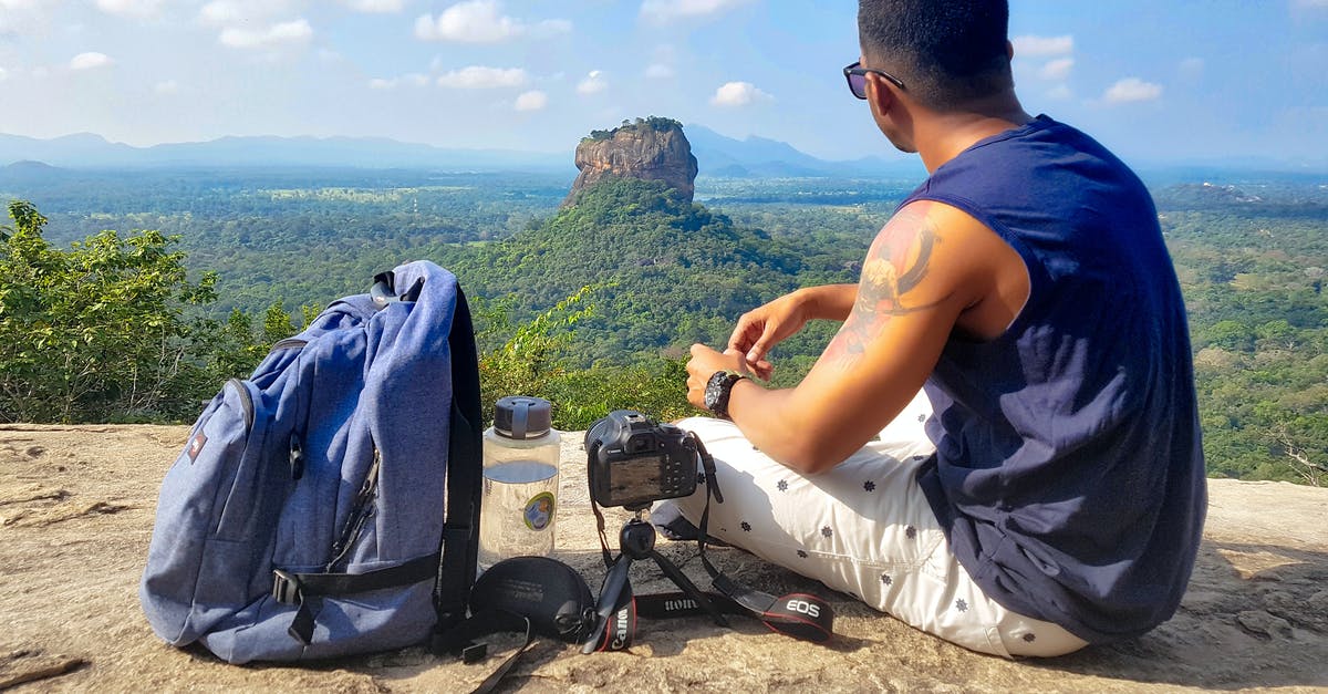 Traveling with BlackDiamond Jetforce avalanche rescue backpack - Man Sitting on Top of Gray Cliff Mountain Beside Backpack, Water Bottle, and Camera