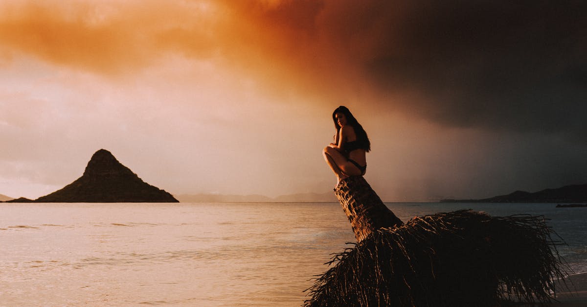 Travelling to Poland alone at the age of 17 - Full body side view of lonely female in swimwear sitting on old buoy near calm ocean against bright cloudy sky at sunset