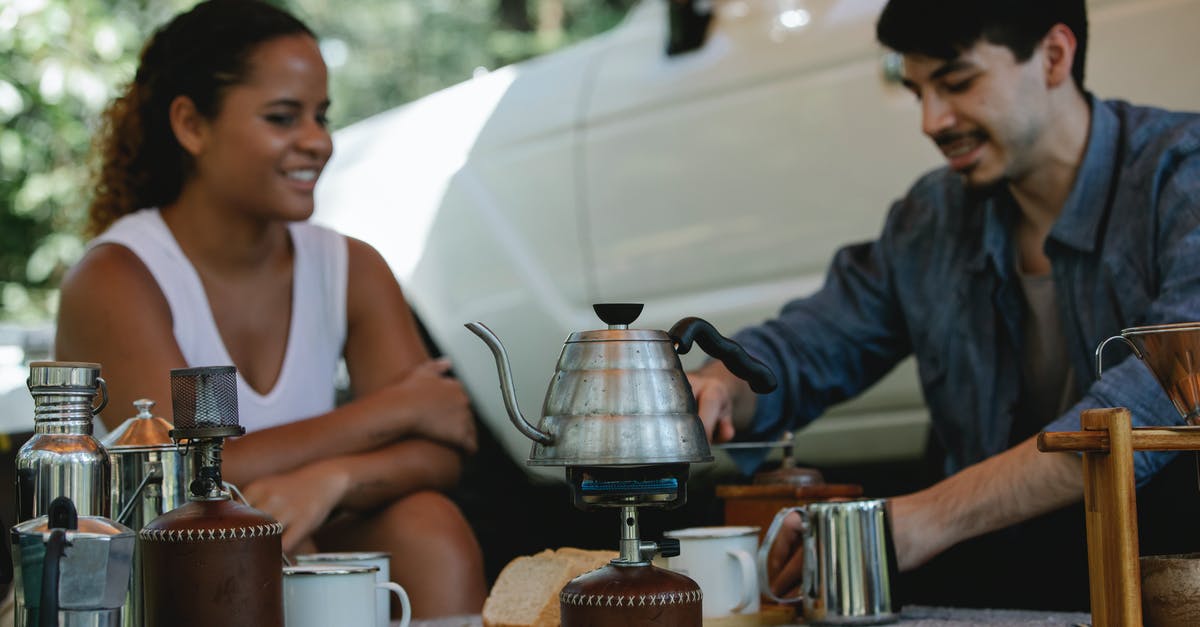 Travelling with a portable Stove and fuel canisters - Young cheerful couple in casual wear boiling water in kettle on gas stove and grinding beans for coffee brewing while spending sunny day in camp