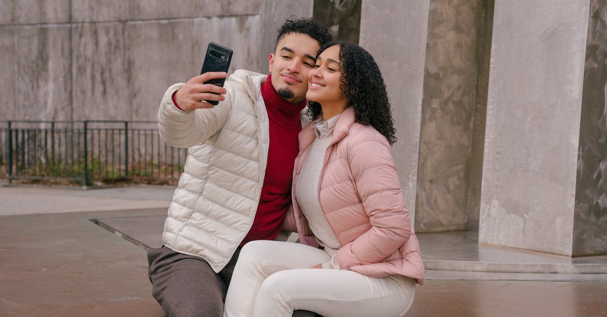 Uk Tier 5 Youth Mobility Visa arrival dates - Delighted young ethnic couple touching cheeks while taking selfie sitting on parapet