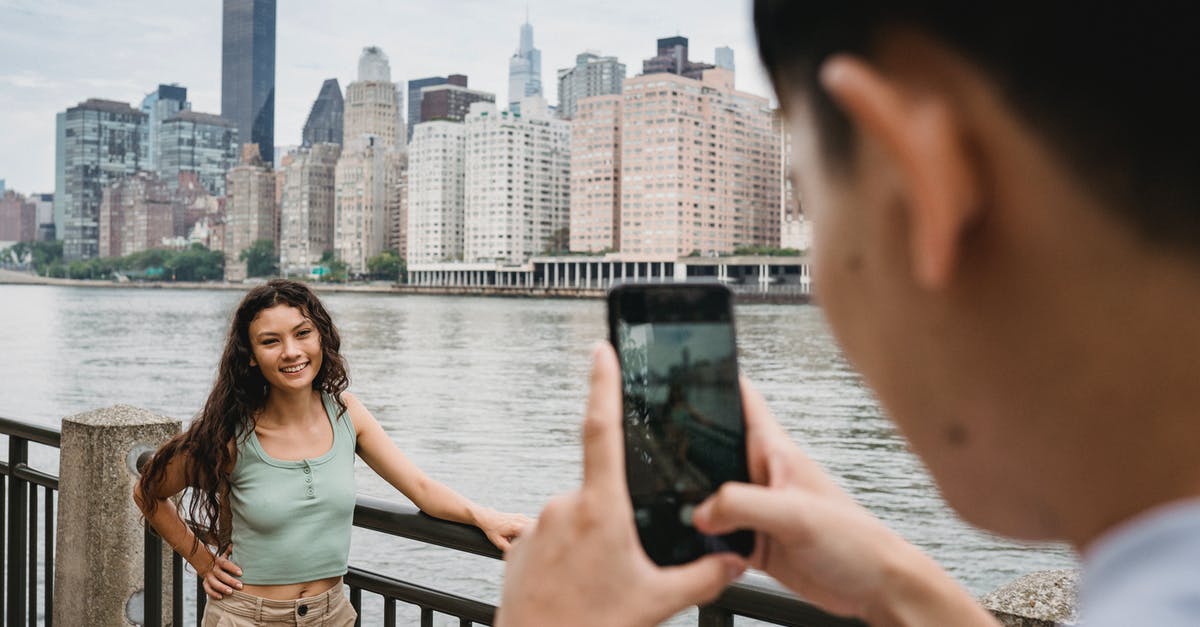 US citizen making money online when in the UK as a tourist - Crop unrecognizable young guy using smartphone while photographing happy young ethnic girlfriend standing on river embankment with hand on waist during trip in New York City