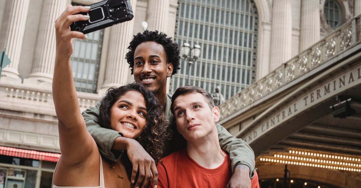 US Customs and connection time - Positive multiethnic friends taking selfie on modern smartphone near railway station building