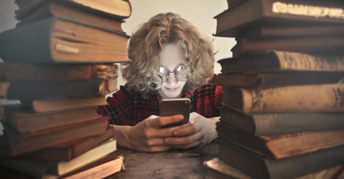 Using library in Frankfurt as a short-term visitor - Focused male student in casual wear and glasses browsing smartphone while sitting at wooden table with stacked old books