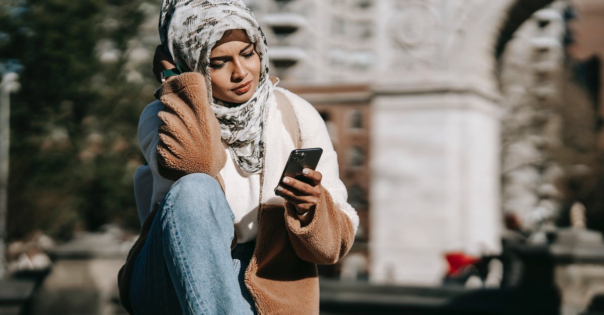 Using Modern Standard Arabic in Arabic-speaking countries - Stylish young Arab female in casual warm outfit and traditional hijab resting on city square and messaging on mobile phone on sunny day