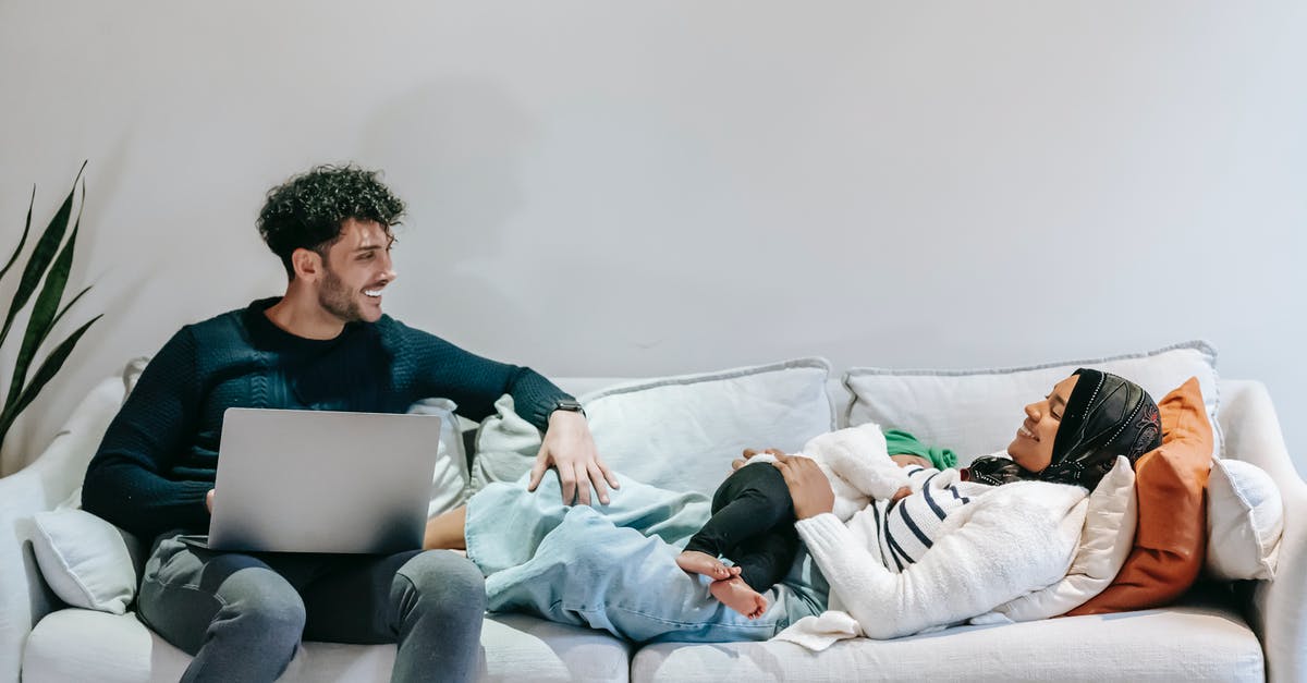 Using no-show ticket for infant - Cheerful young black Muslim woman in casual clothes and hijab relaxing on bed with cute infant in arms and communicating with ethnic husband working remotely on laptop