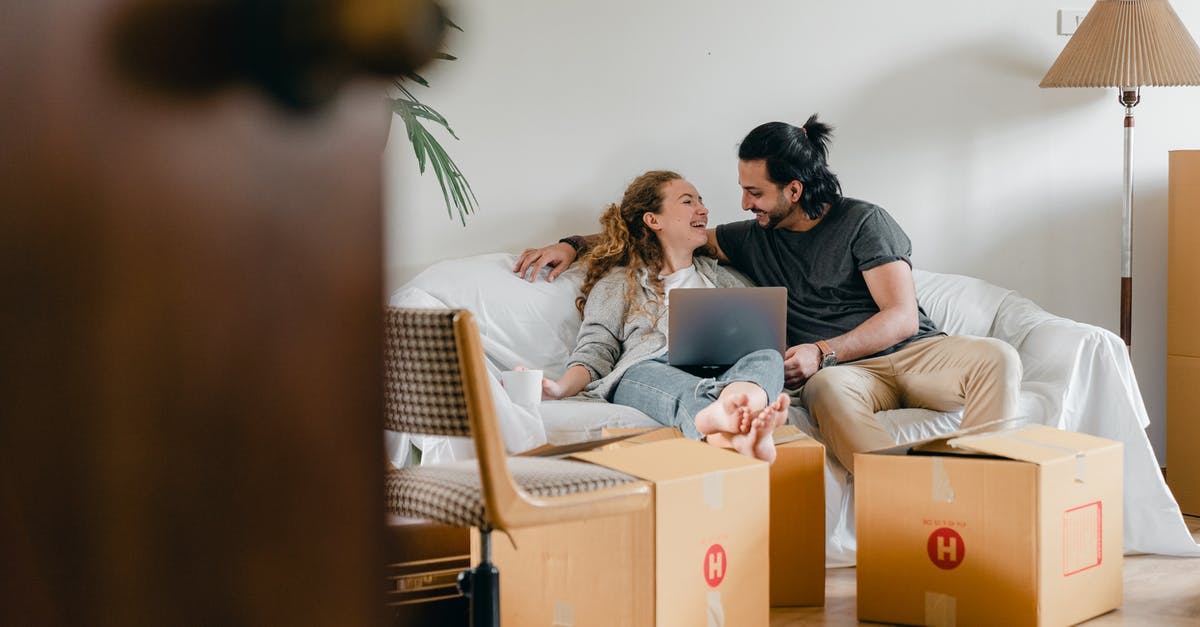 Using parent's property and will as evidence of assets - Cheerful couple in casual clothes sitting on comfortable sofa with laptop and looking at each other while moving to new apartment