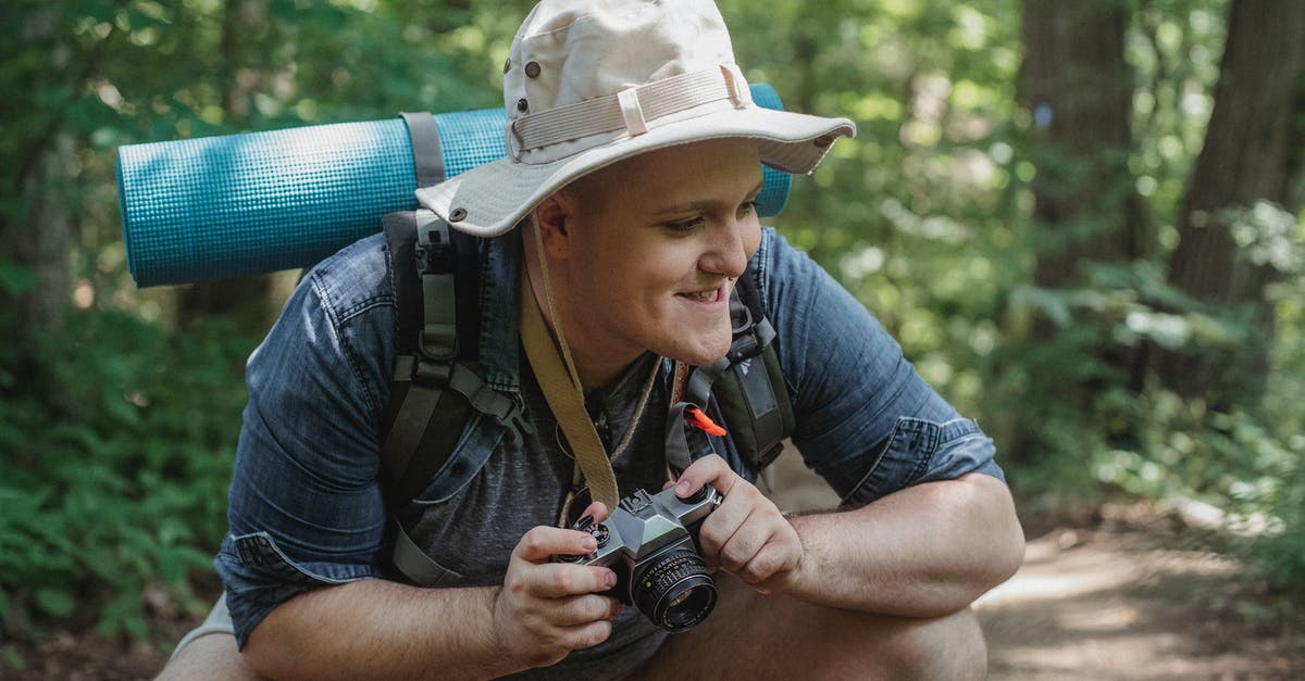 Using squat toilets with arthritic knees - Young smiling male hiker with mat and photo camera squatting on pathway with trees behind while looking away in woods