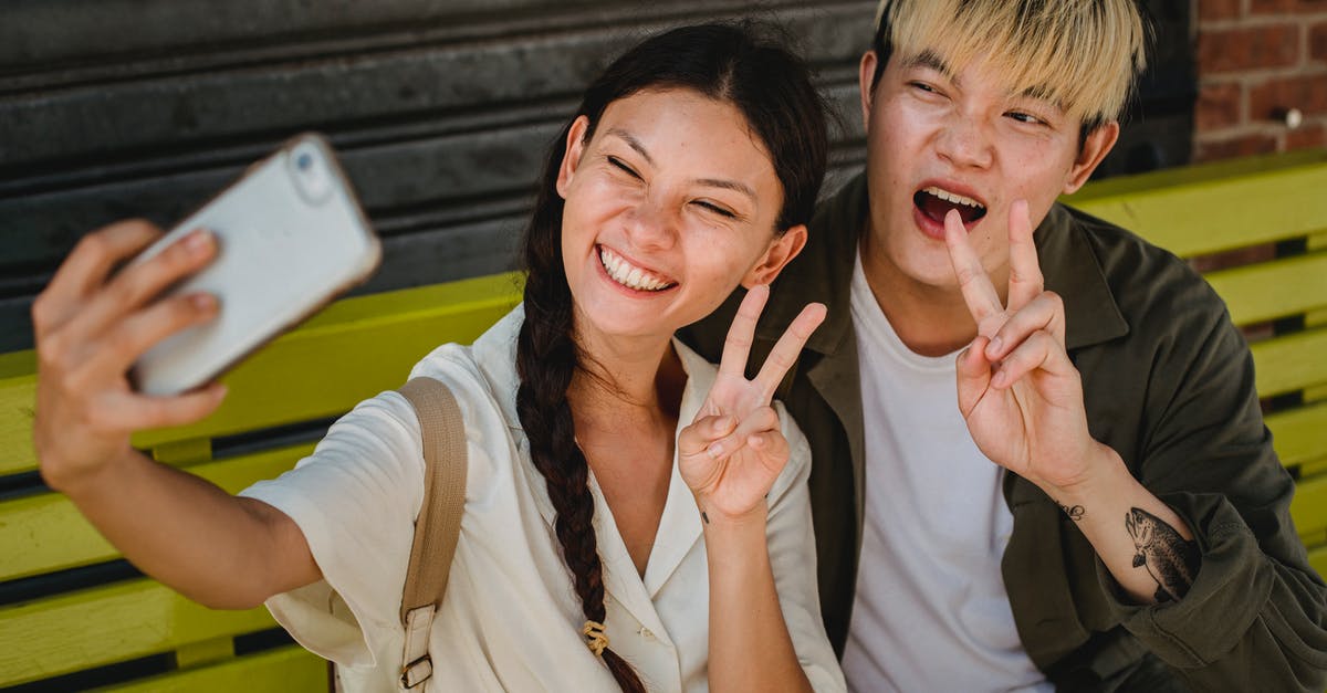 Using two diferent tickets and airlines - Delighted young Asian couple wearing casual clothes taking selfie on modern mobile phone and showing v sign while sitting on street bench