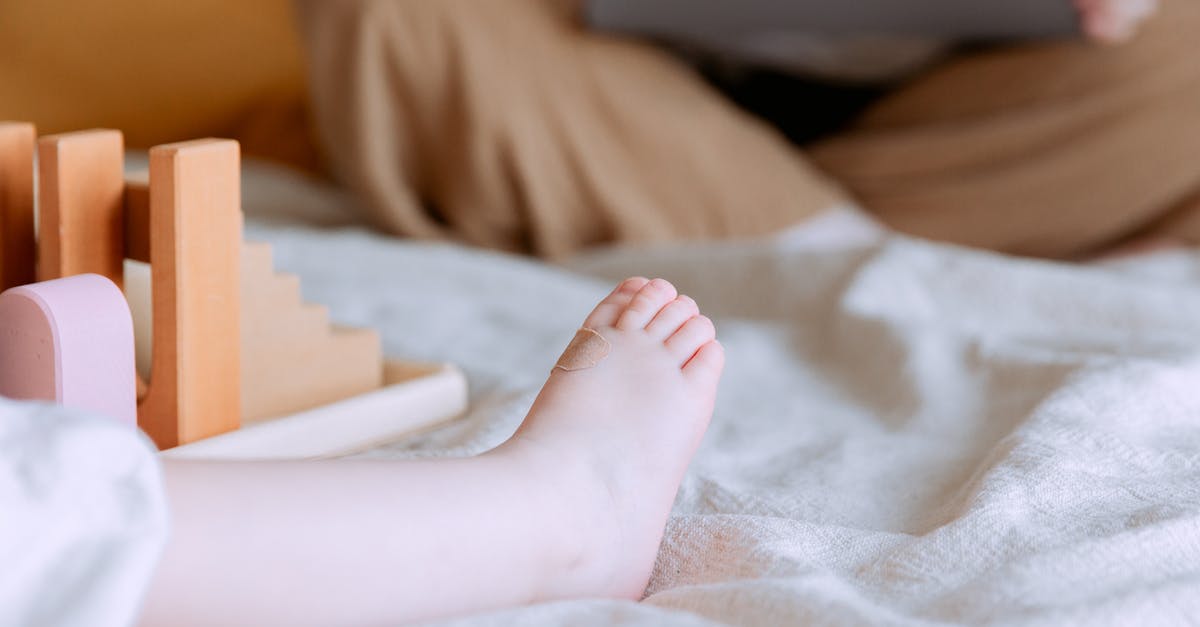 Vancouver YVR getting to quarantine with toddler - Crop faceless toddler sitting on bed with colorful wooden building blocks while mother sitting with legs crossed doing remote work with laptop on knees on background