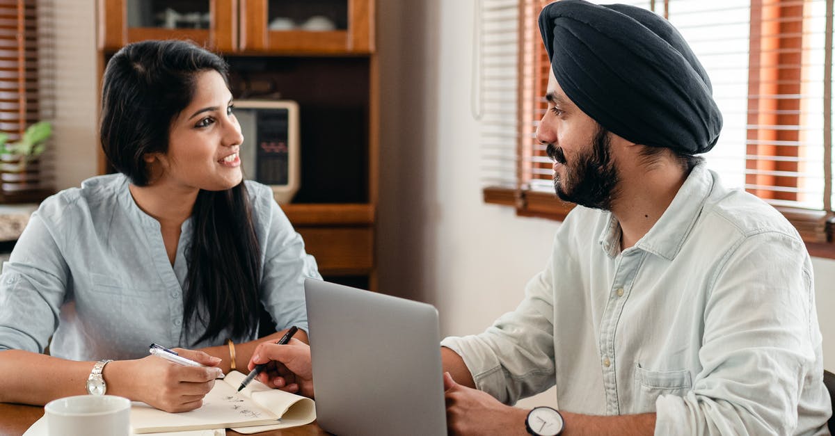 Visit to Switzerland by an Indian couple - Indian male in turban and wristwatch typing on netbook while interacting with happy female coworker with notebook and pen sitting at table in office and looking at each other