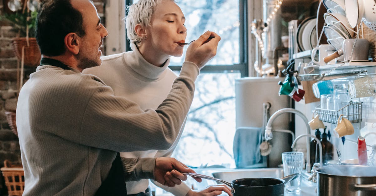 Visiting my spouse in UK [closed] - Side view of content ethnic man in casual clothes and apron feeding happy wife while preparing delicious dinner at stove in kitchen