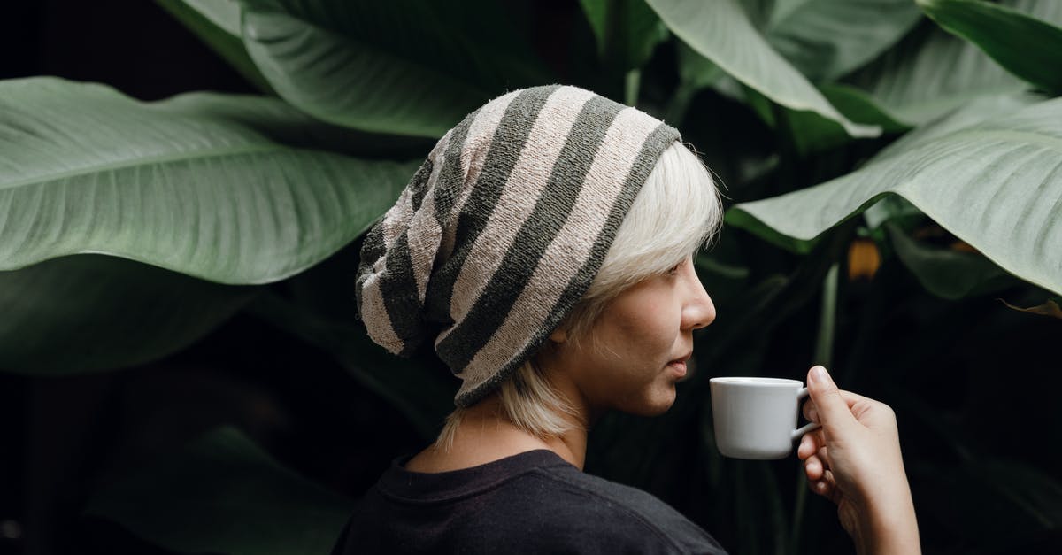 Visiting the Edinburgh Fringe (events information) - Side view of young lady in striped slouchy beanie and black tee with blond fringe drinking cup of aromatic coffee standing near big house plant with large green leaves