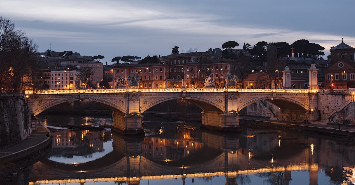 Websites to buy complex air tickets - Aged masonry bridge with arches over Tiber River against urban house facades under cloudy sky in evening Rome Italy