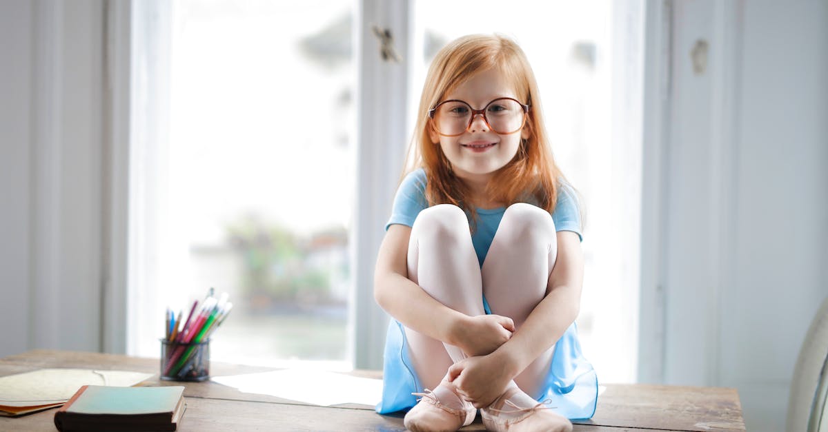What are good holidays for a disabled adult and a small child? - Joyful red haired schoolgirl in blue dress and ballet shoes smiling at camera while sitting on rustic wooden table hugging knees beside school supplies against big window at home